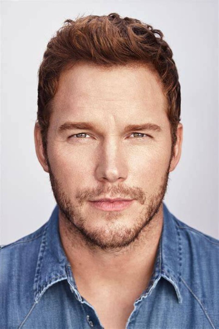 Chris Pratt smiling in front of a dark gray background with repeating gold logos