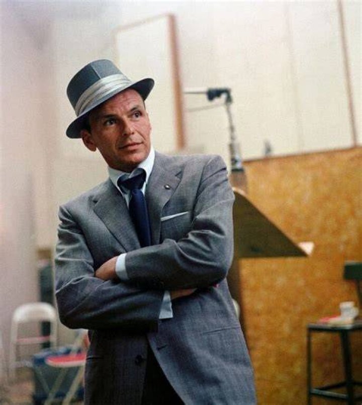 Frank Sinatra wears a suit and fedora and sits against a blue background.