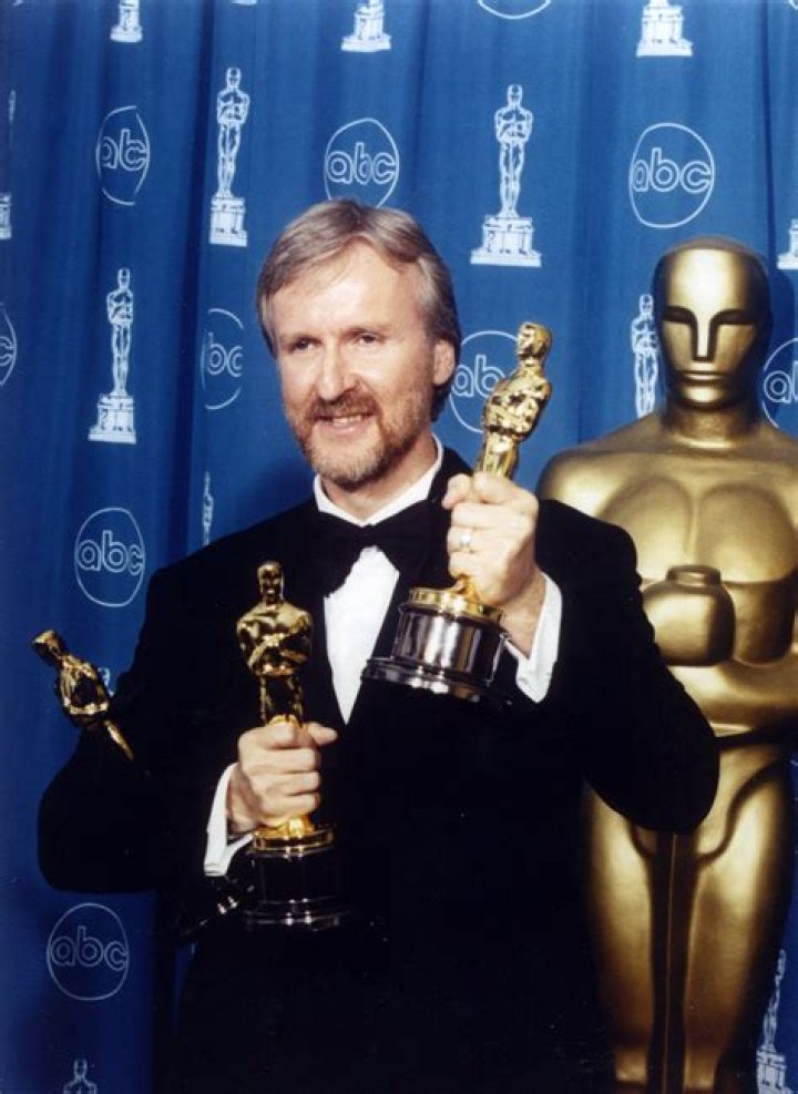 James Cameron, holding two Oscars, which he almost used to hit Harvey Weinstein. He's wearing a tux, standing in front of a blue background and a large version of the Oscar statuette.
