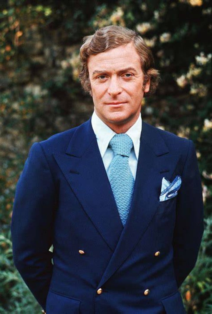 Michael Caine wears a suit jacket and stands outside.