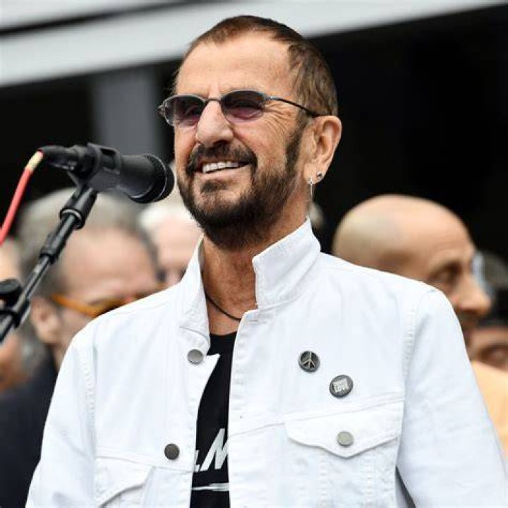Ringo Starr holds a microphone and lifts his arms up to show peace signs while standing onstage.
